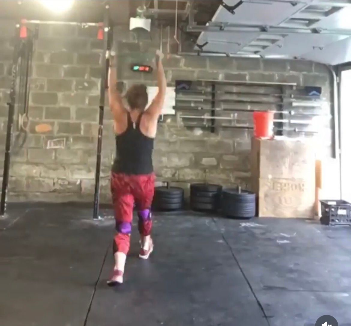 Person lifting a barbell overhead in a gym, facing away, with stacked weights and a red bucket nearby