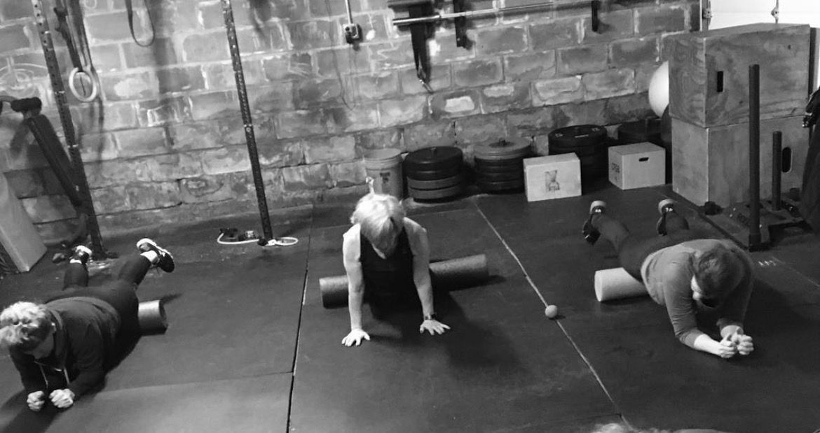 People doing plank exercises on mats in a gym, with kettlebells and brick walls in the background