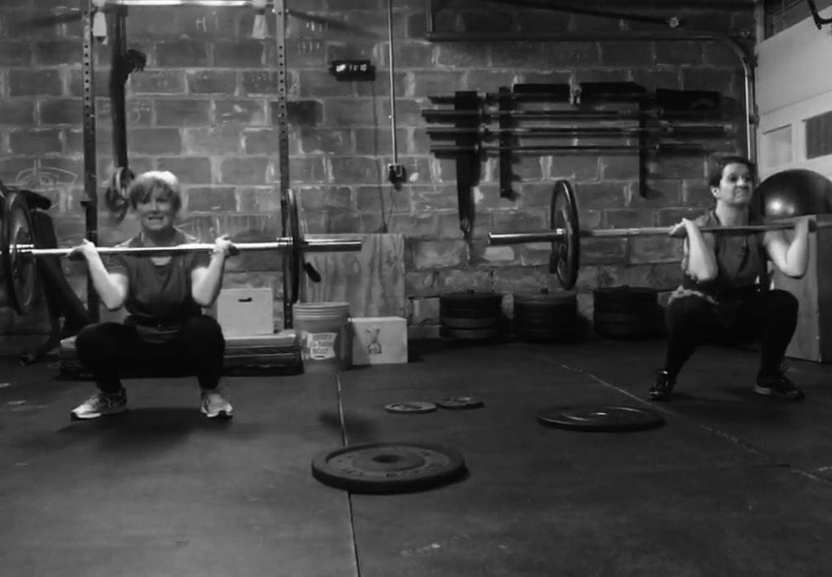 Two people squatting with barbells in a gym, black-and-white photo.