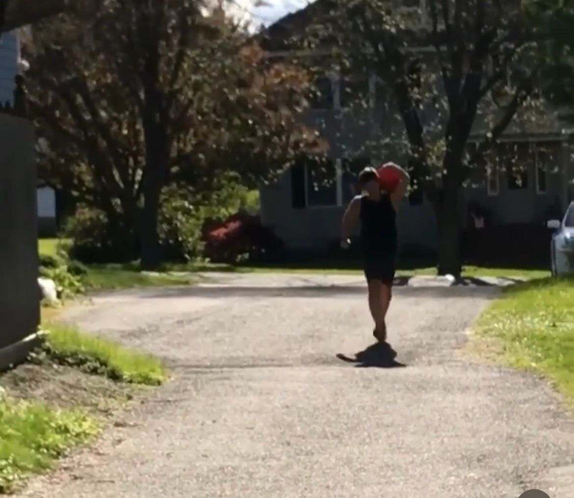 Person standing on a sunlit driveway holding a red ball, with a house and trees in the background.
