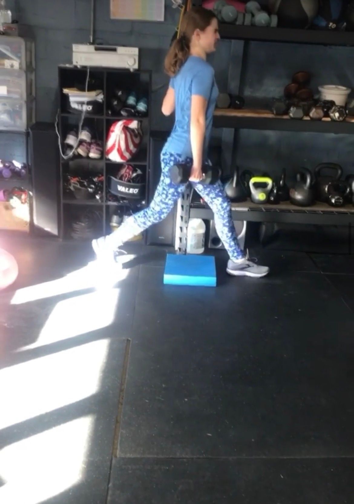 Woman performing a split stance on a blue block in a gym, wearing a blue shirt and patterned leggings.