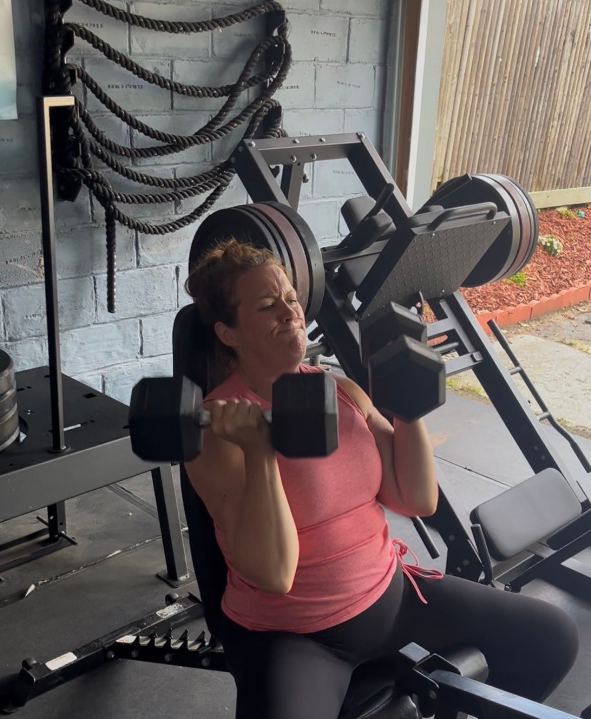 Person doing seated dumbbell shoulder press in a gym, wearing a pink top.