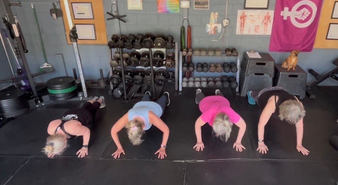 Four people doing push-ups on gym mats in a workout studio, with dumbbells and flags in the background.