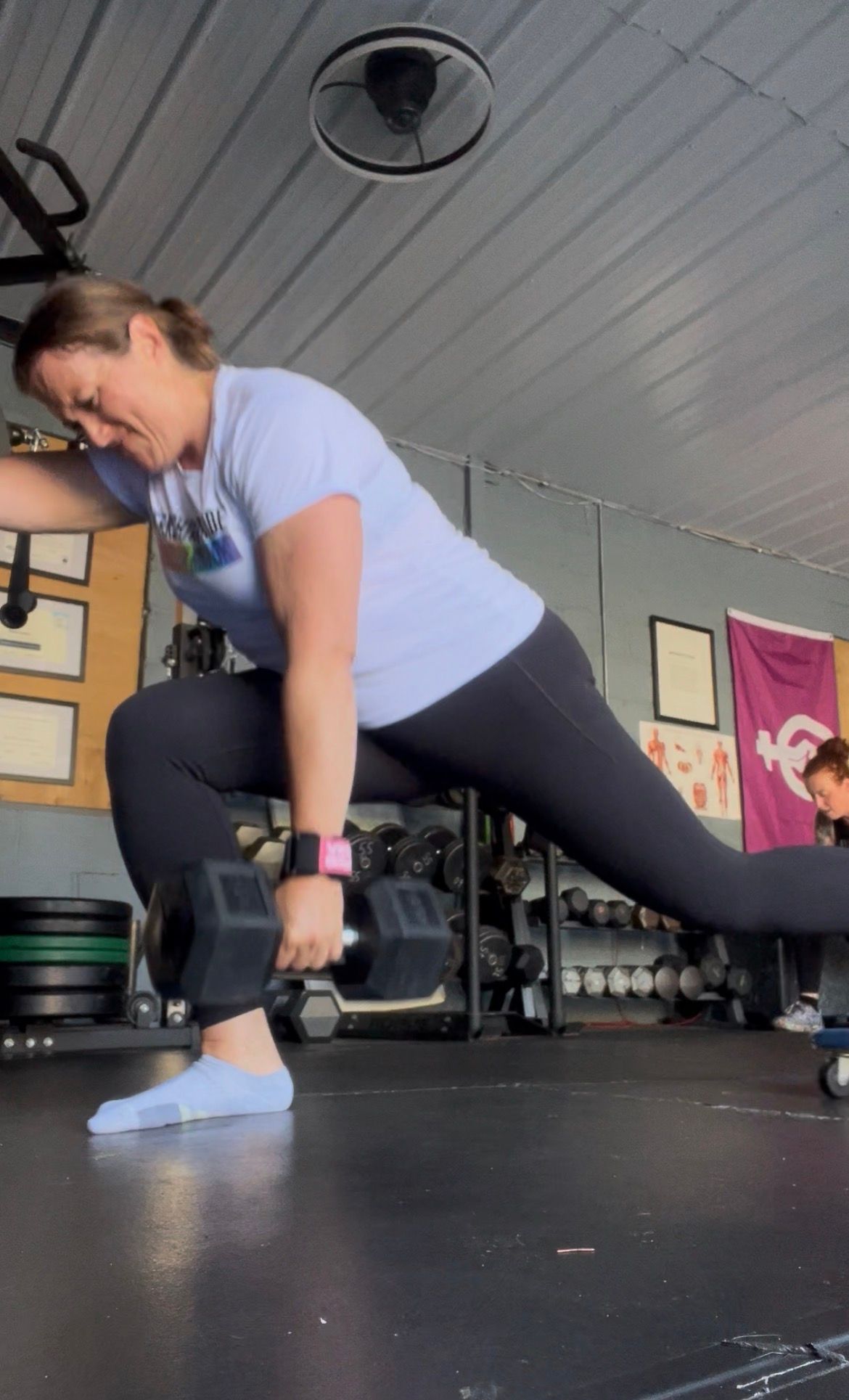 Person lunging with dumbbells in a gym, wearing gray shirt and black leggings.