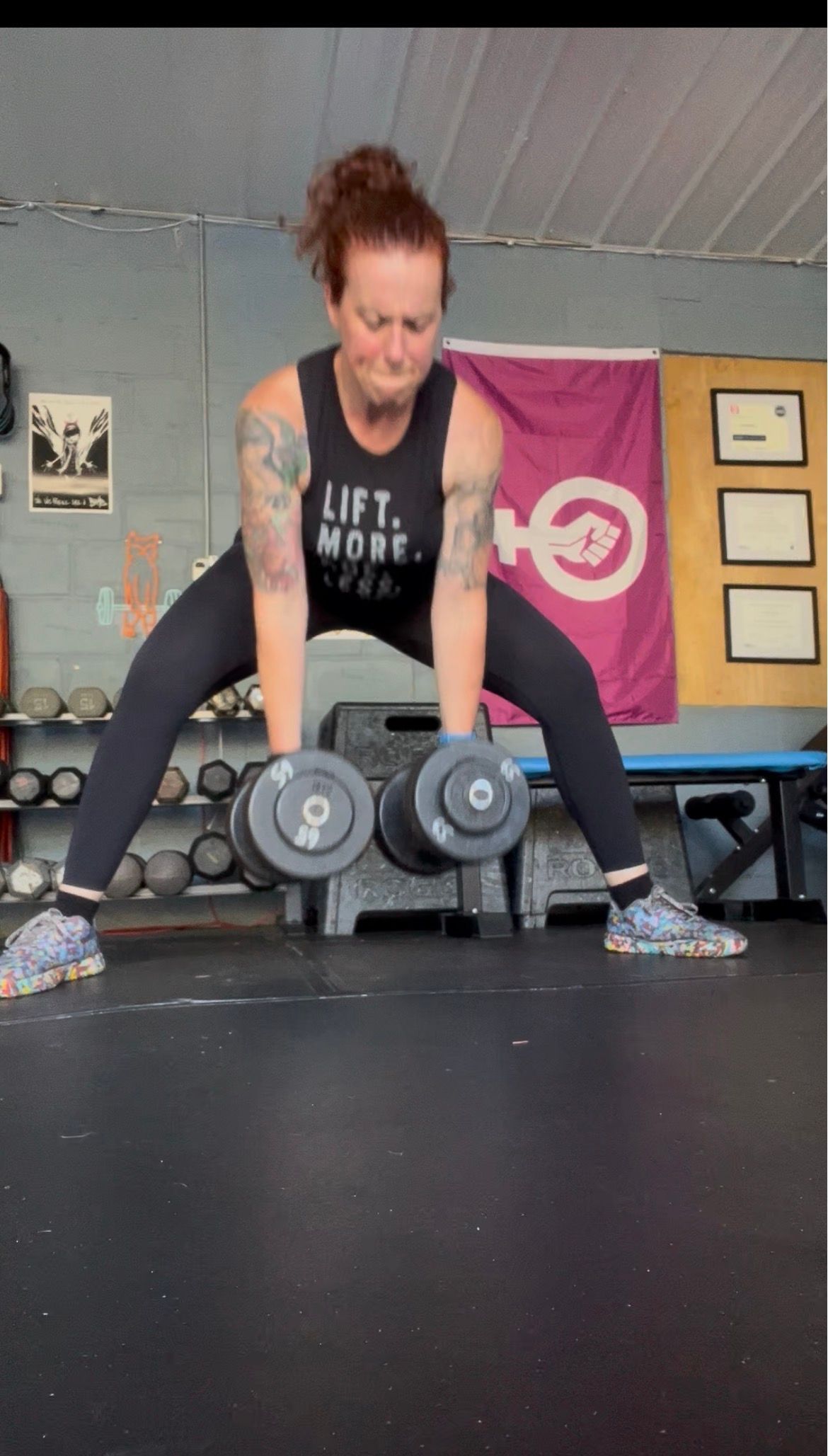 Person deadlifts dumbbells in a gym, wearing a black tank top and leggings, with a flag on the wall.