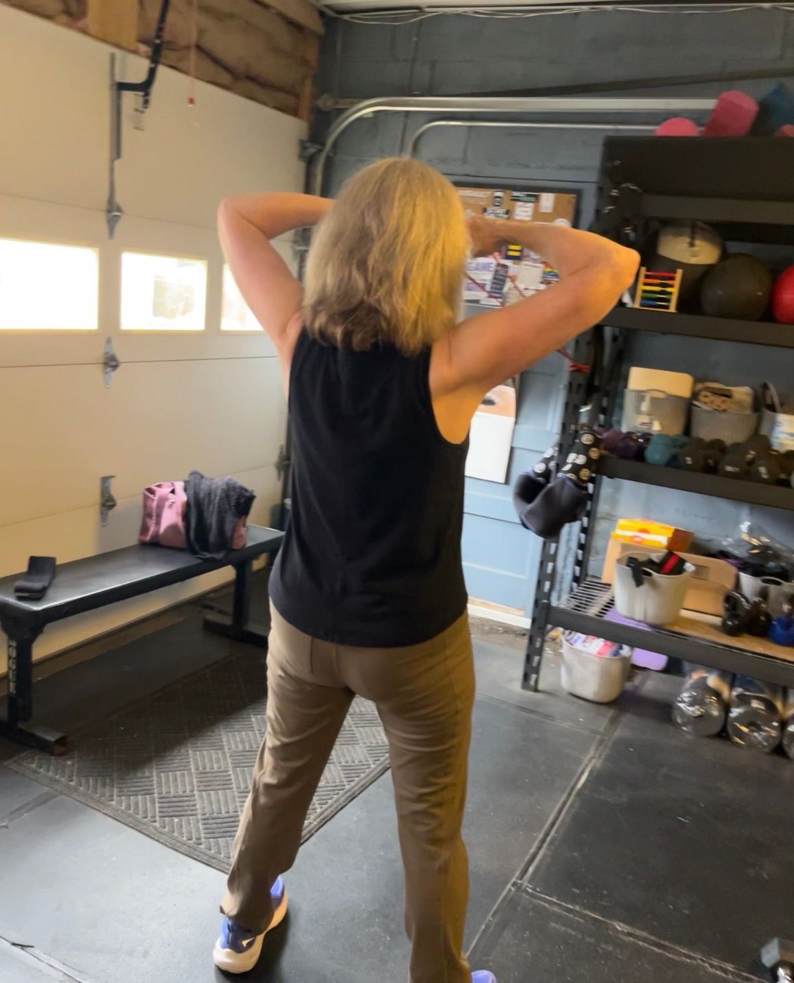 Person standing in a garage gym, facing away, with hands behind head near shelves of weights and equipment