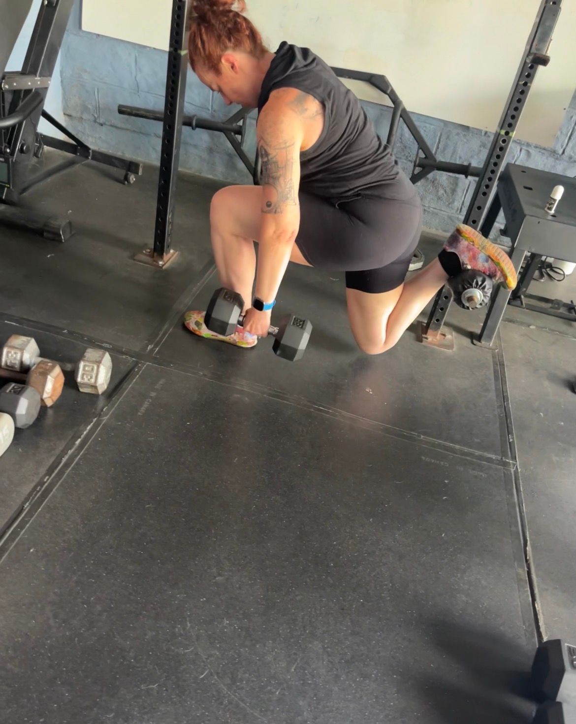 Woman doing a dumbbell row in a gym, wearing dark workout clothes near scattered weights.