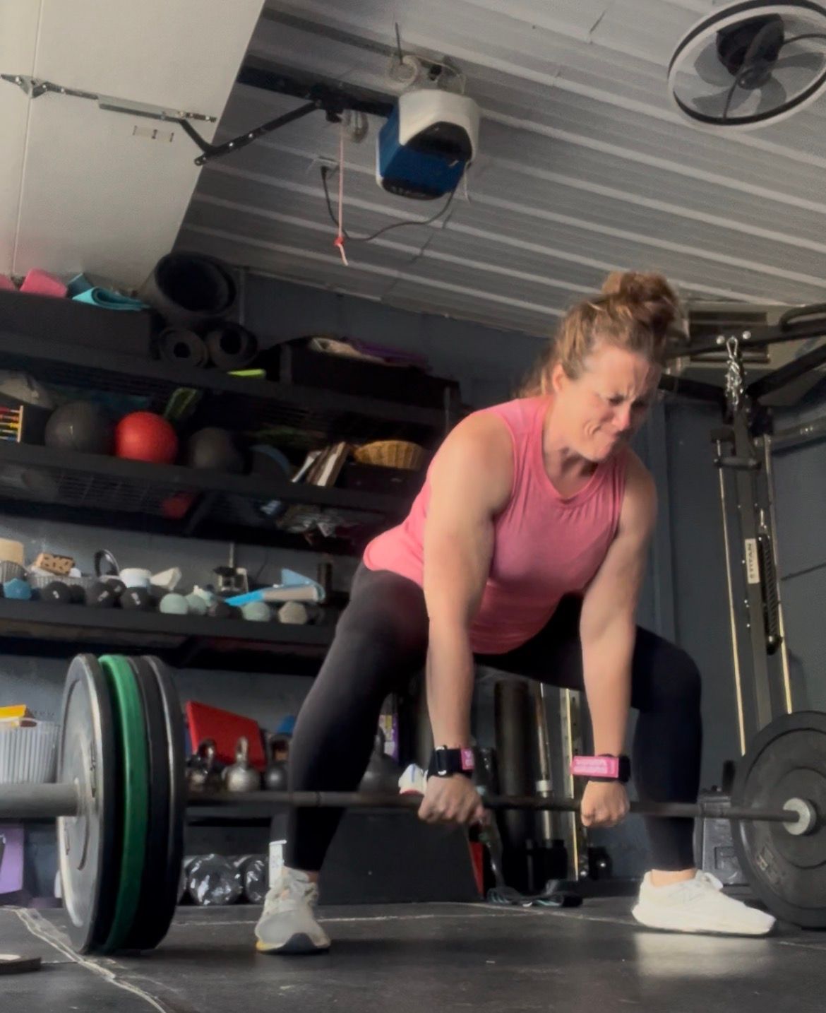 Person deadlifting a barbell in a garage gym, wearing a pink top and black leggings.