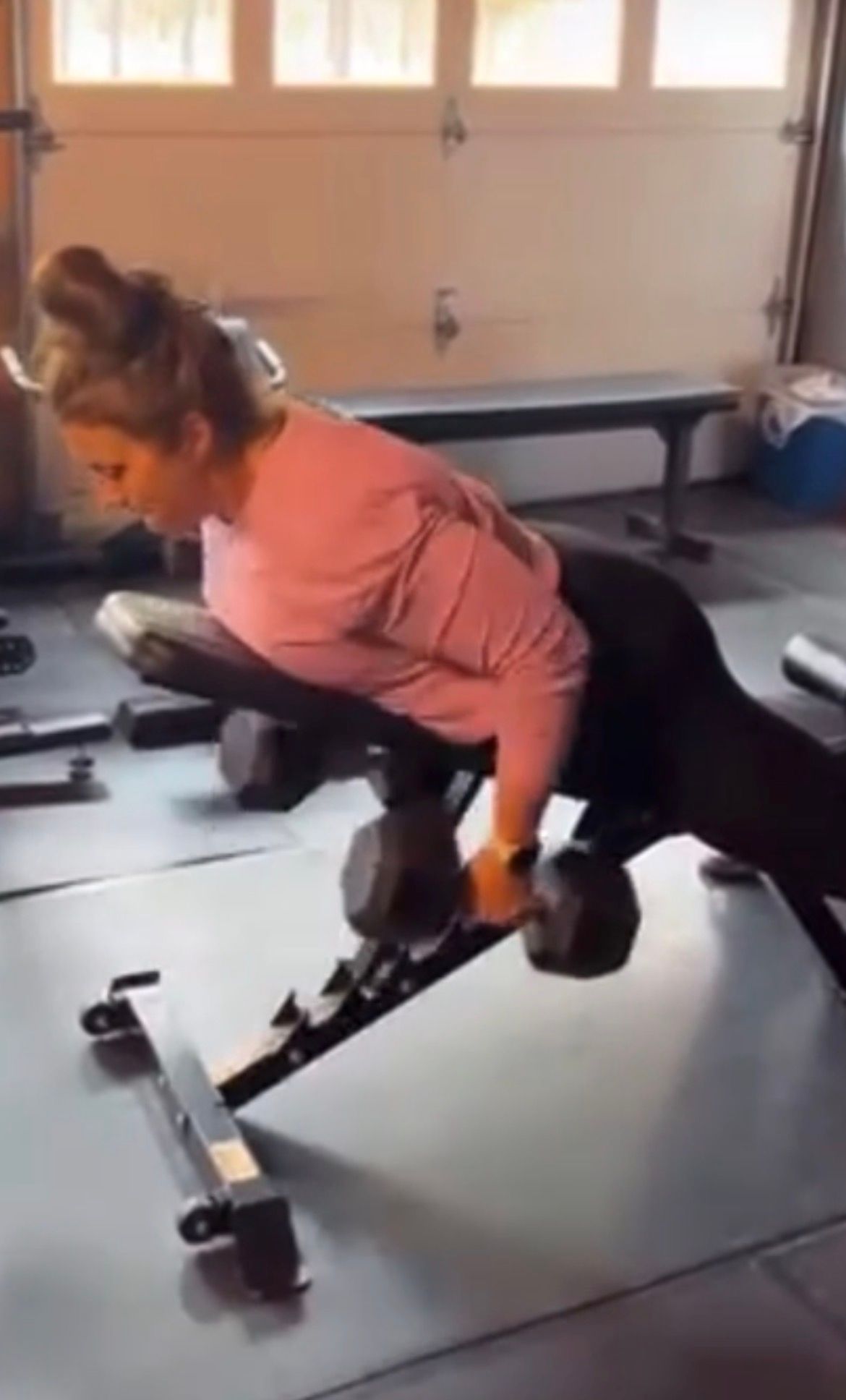Woman doing a dumbbell row on a bench in a gym, wearing a pink top and black leggings.