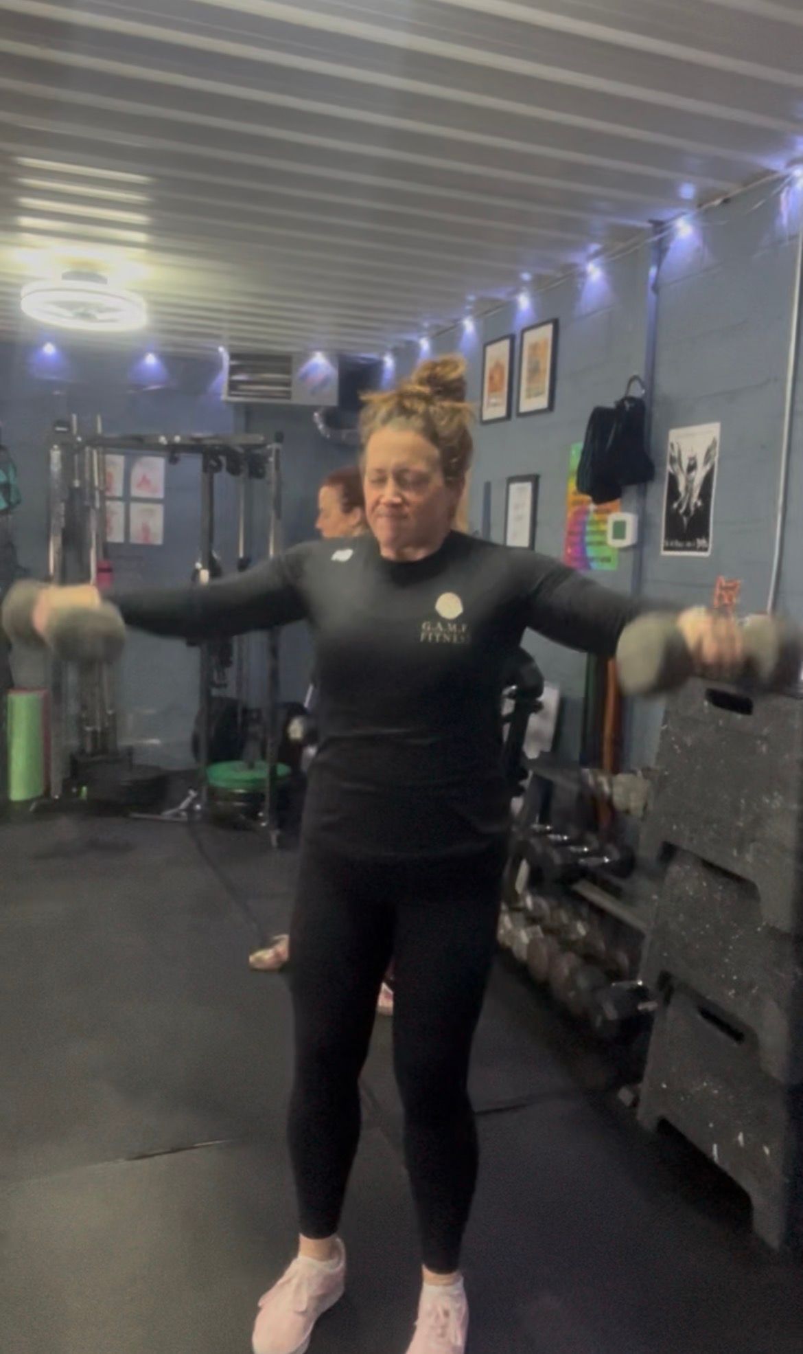 Woman exercising with dumbbells in a gym, arms raised, wearing black workout clothes.