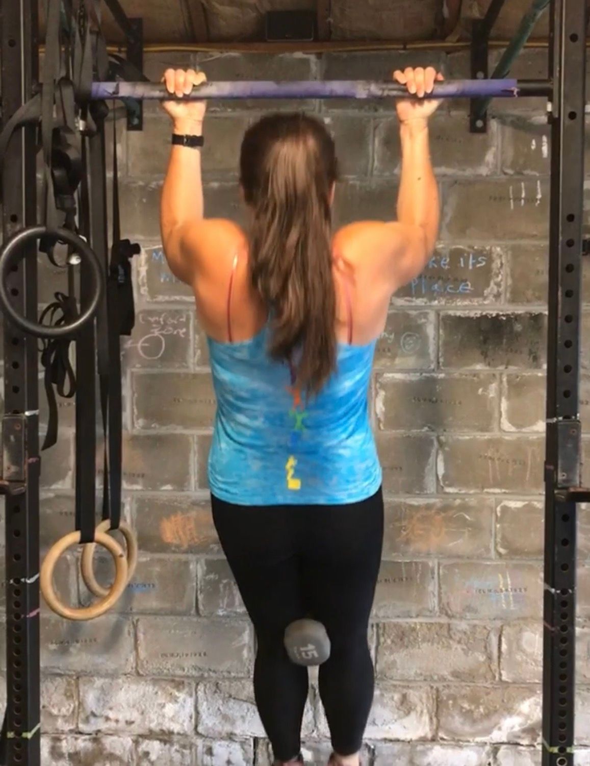 Woman doing a pull-up on a bar in a gym, wearing a blue tank top and black leggings.