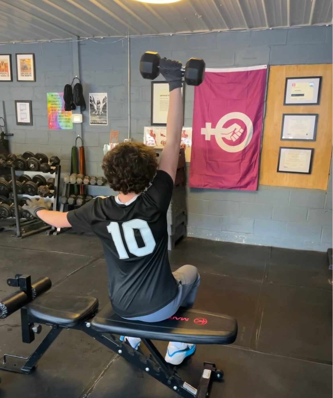 Person seated on a bench presses a dumbbell overhead in a gym, wearing a black jersey with number 10.