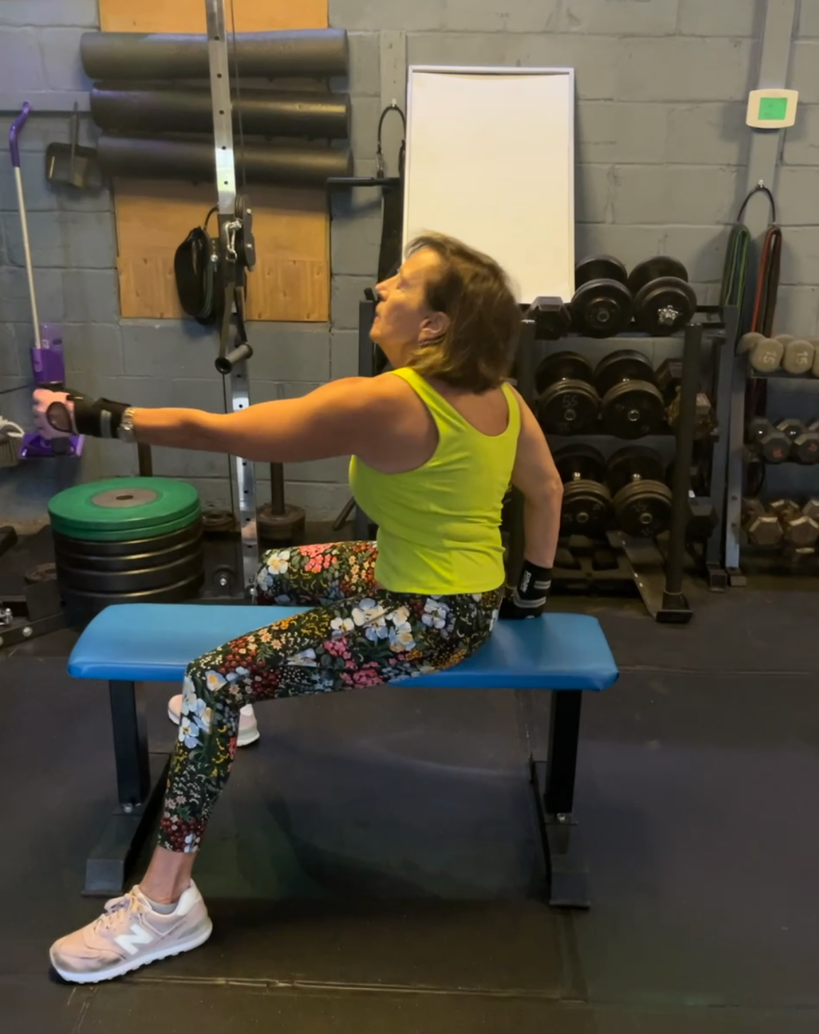Woman in neon workout clothes sitting on a bench, extending one arm in a gym.