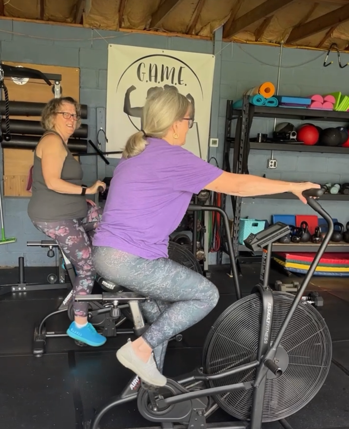 Two people exercising on stationary bikes in a gym, one in a purple shirt reaching forward.