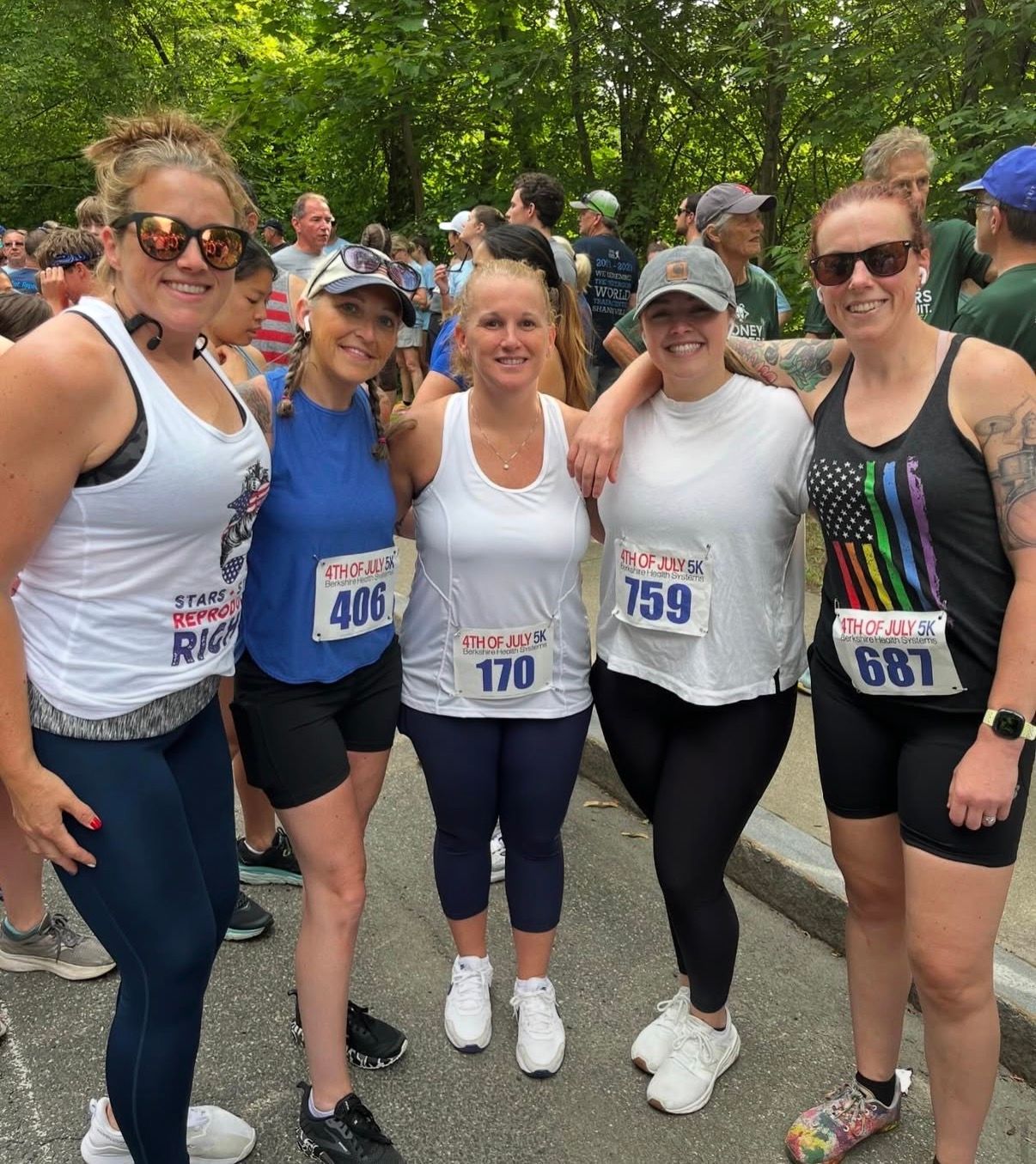 Five runners posing together outdoors in a wooded park before a race, smiling and wearing bibs.