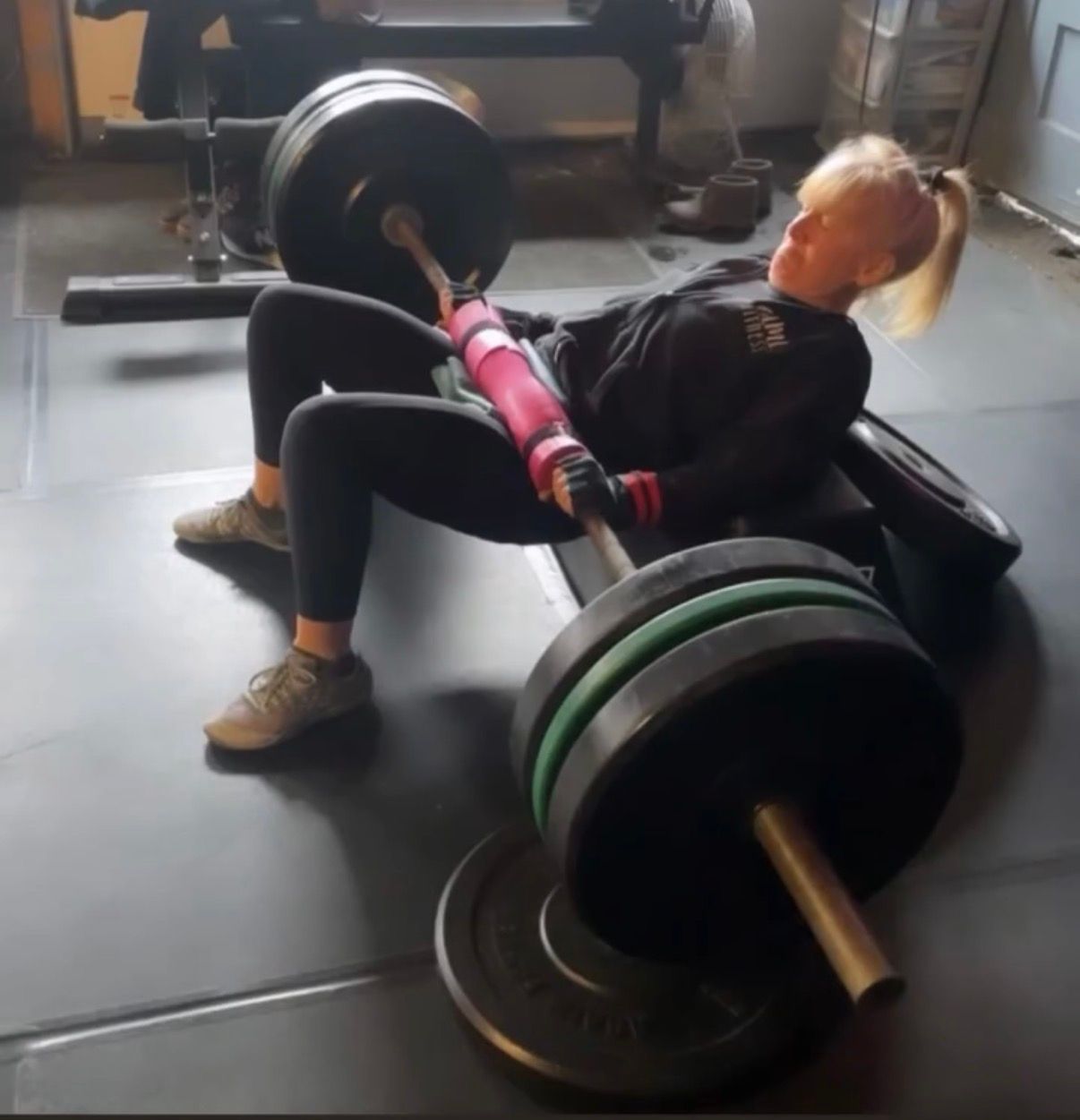 Person doing a barbell hip thrust on a gym floor with a pink pad and heavy weights