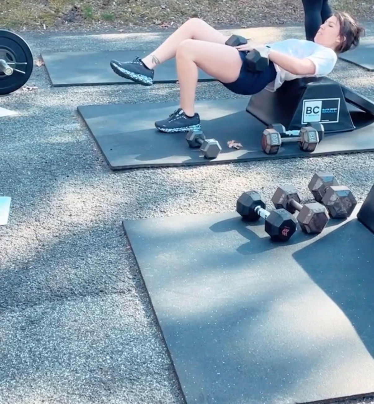 Person doing a floor hip bridge workout on mats beside dumbbells outdoors.
