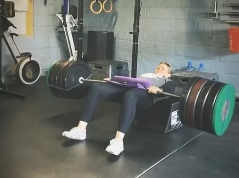 Person performing a barbell hip thrust in a gym, wearing black leggings and white sneakers.