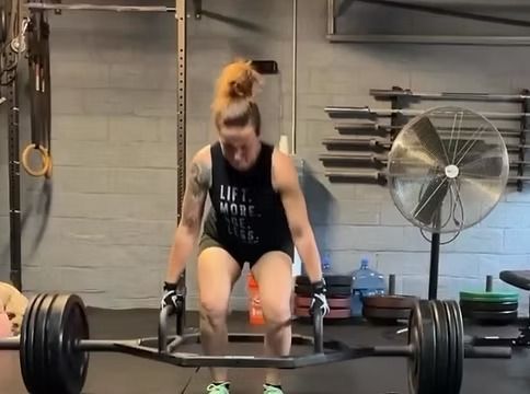 Woman deadlifting a barbell in a gym, wearing black workout clothes and orange shoes.