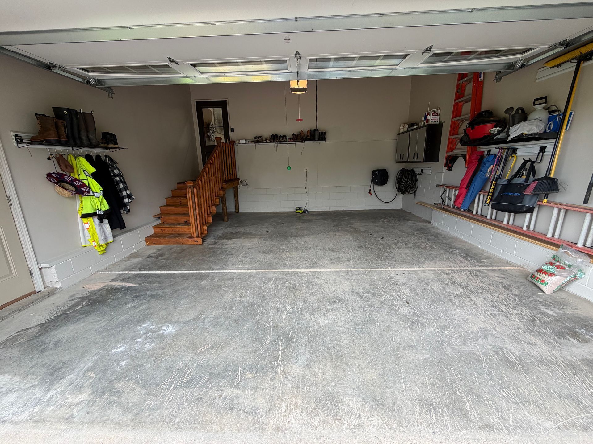 Spacious garage with concrete floor, wall shelves, hanging gear, and a wooden staircase along the left wall