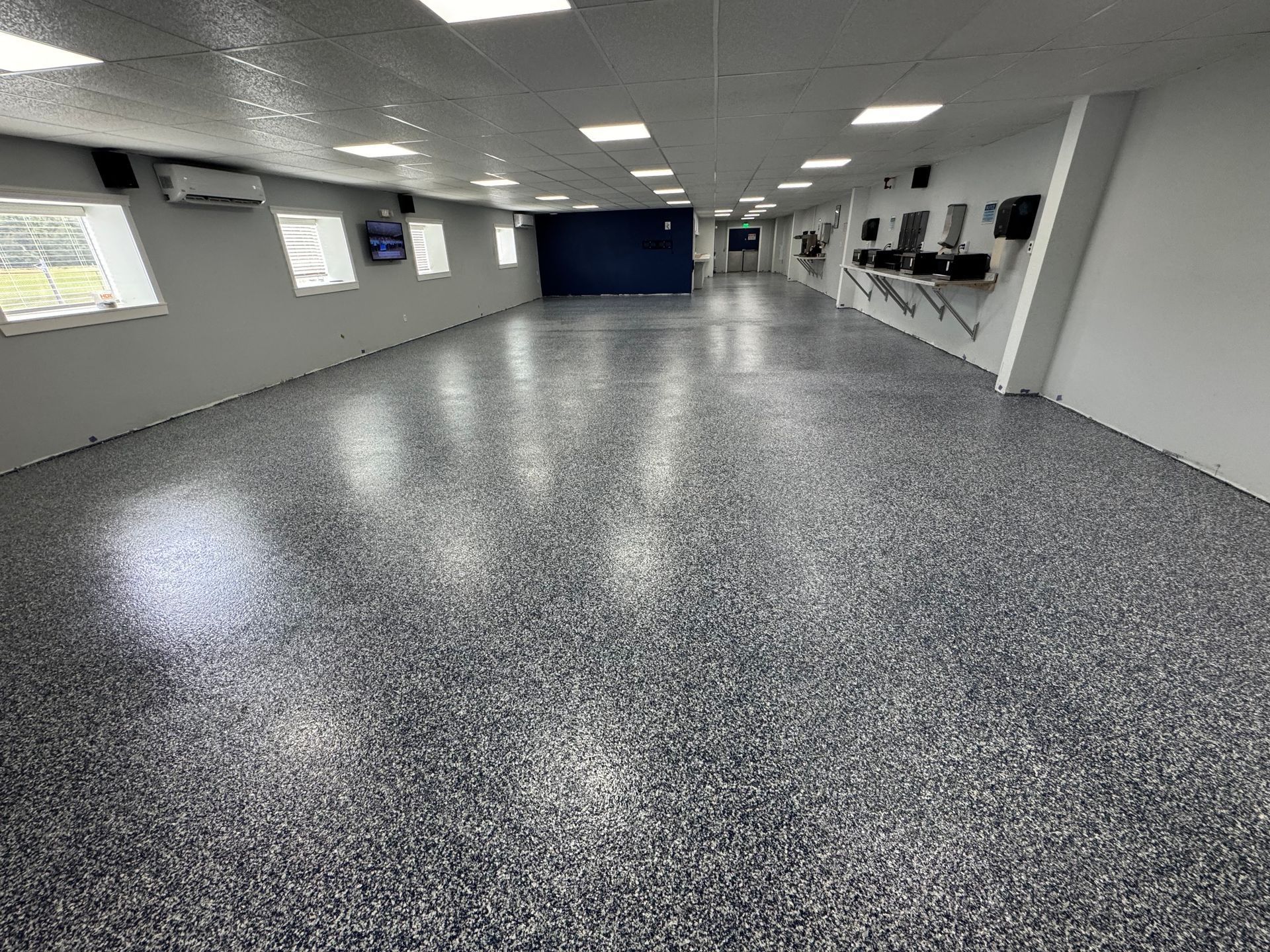Empty indoor room with gray speckled floor, white walls, windows, and wall-mounted screens