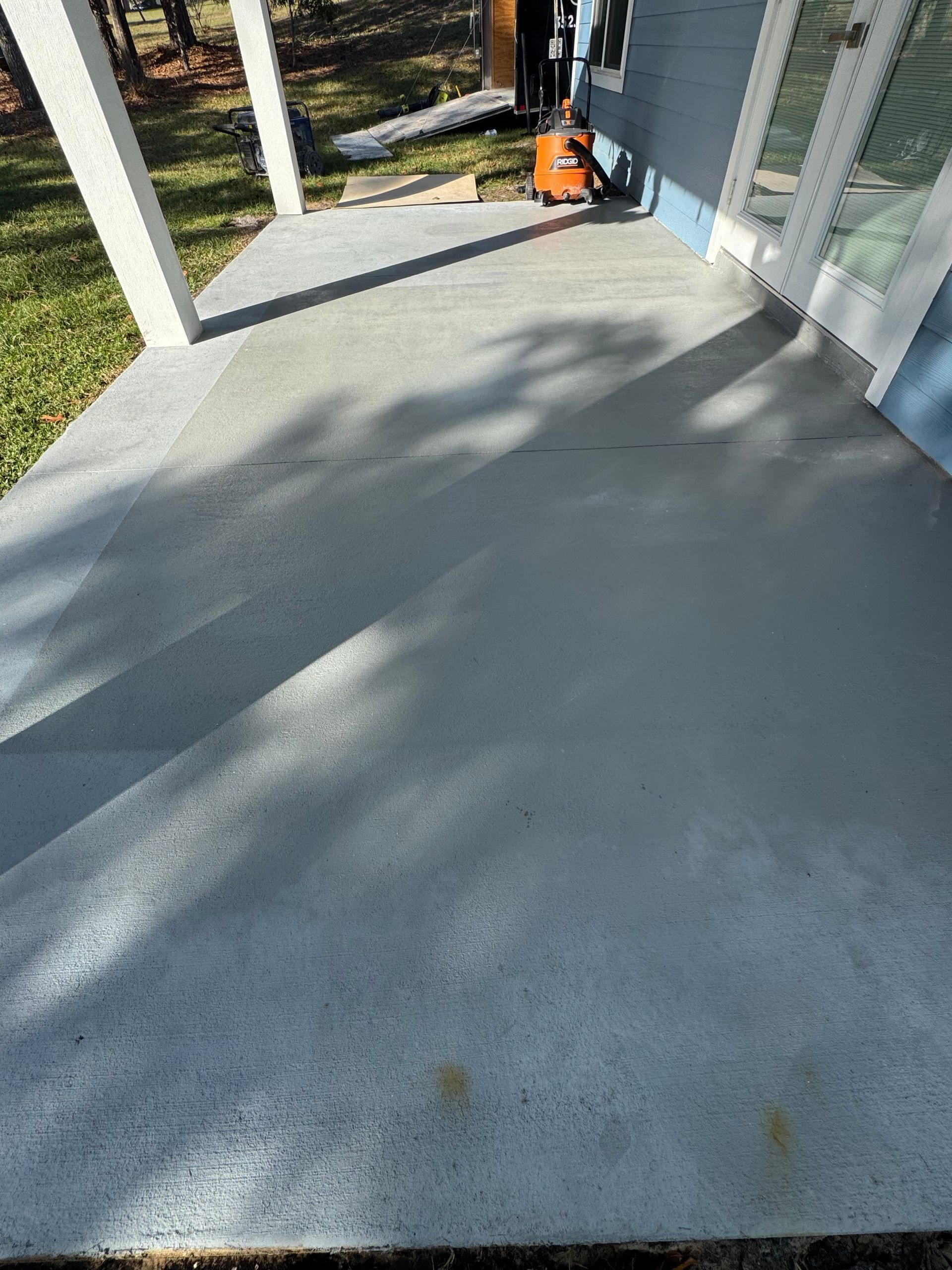 Sunlit concrete porch with white posts, glass doors, and an orange tool near the wall