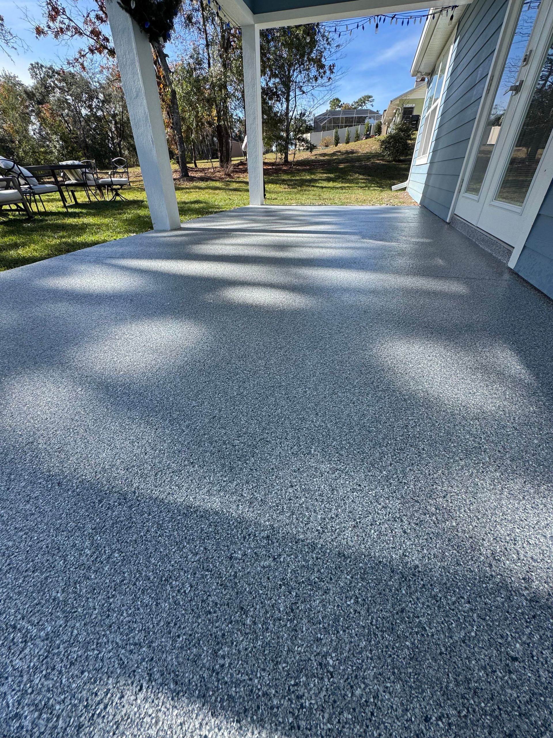 Covered porch with gray speckled floor, white columns, and a view of a sunny yard beyond.