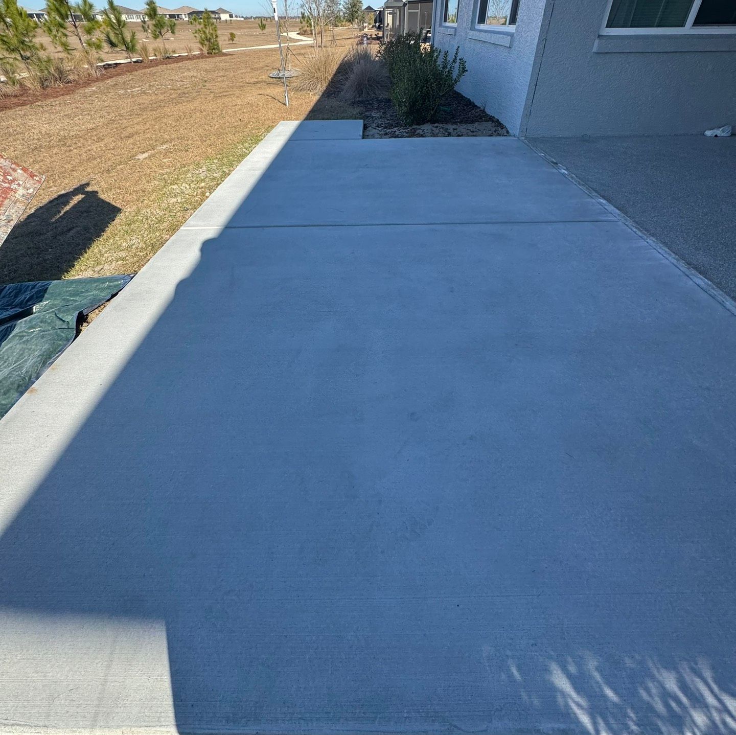 Concrete patio beside a house, partly shaded, with a grassy yard and small trees nearby