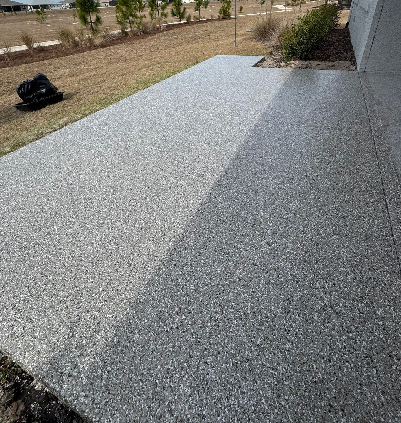 Gravel-covered patio beside a house, with a planted bed and a black bag on the lawn.