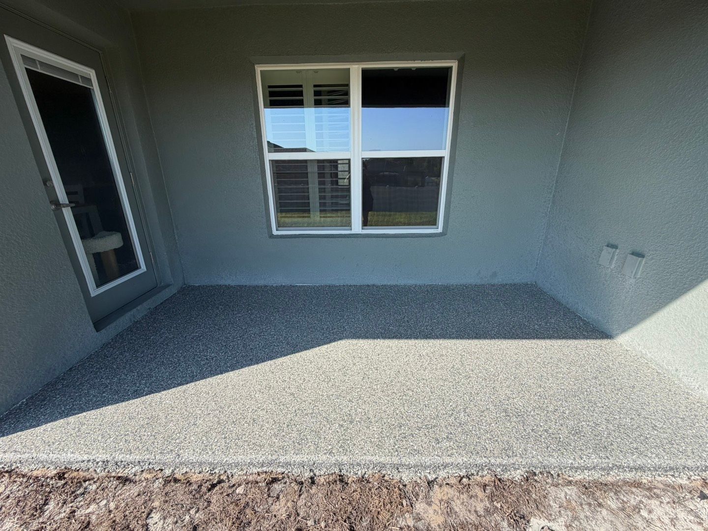 Small covered concrete patio with a window and door, lit by sunlight.