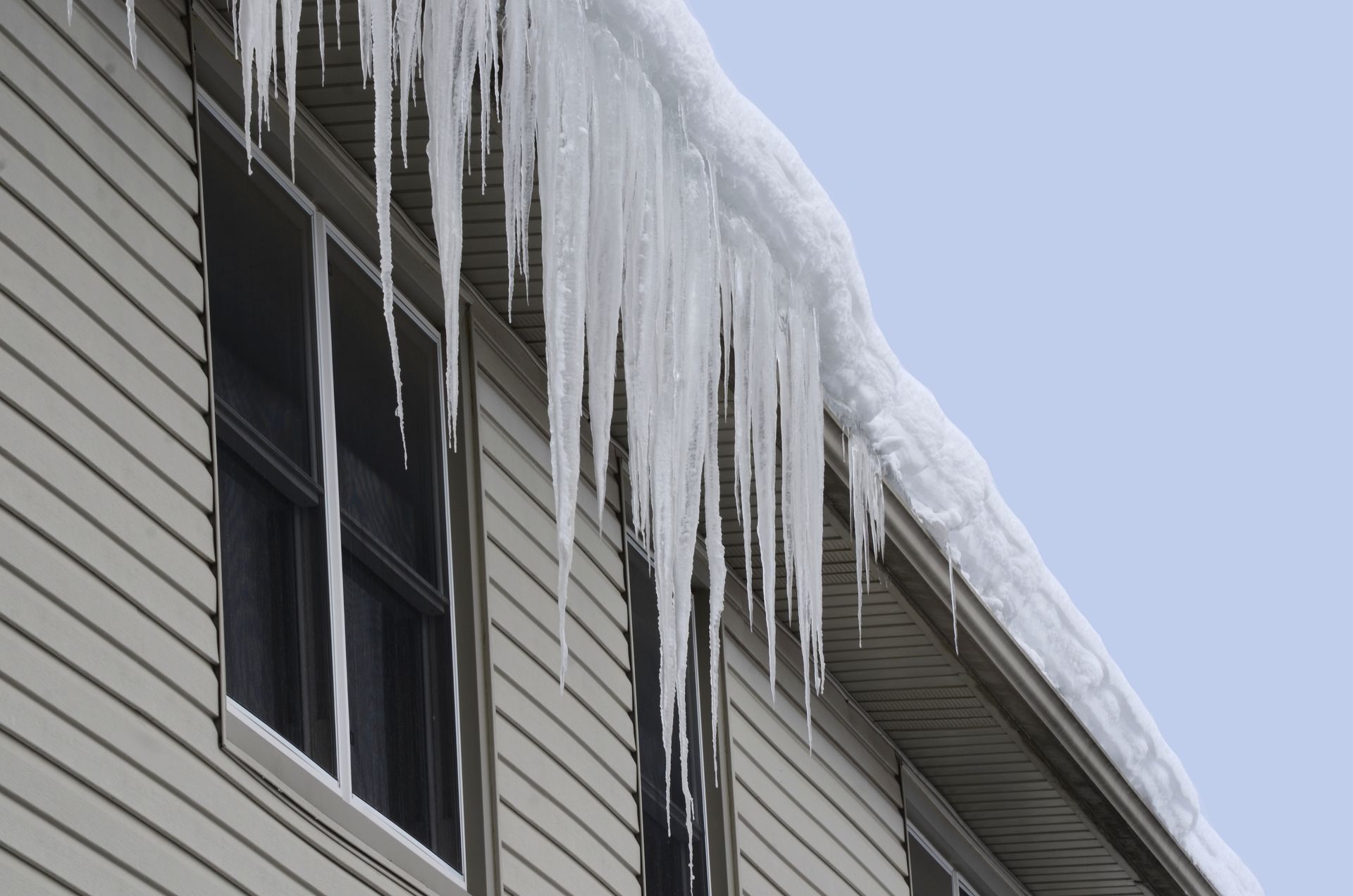 Long icicles hang from a roof on a house with light siding; a clear sky.