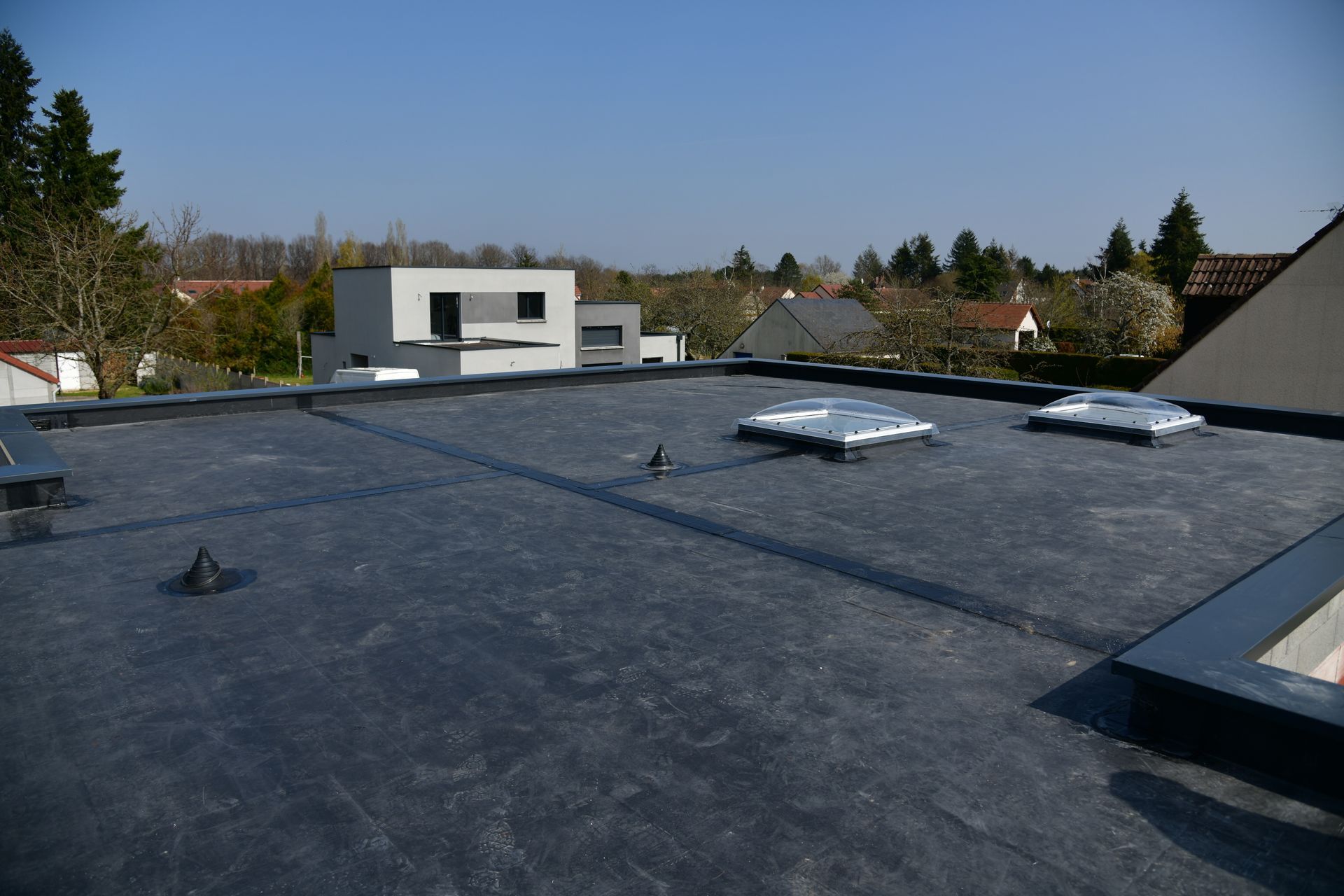 Toit plat noir avec puits de lumière, entouré de maisons et d'arbres par une journée ensoleillée.