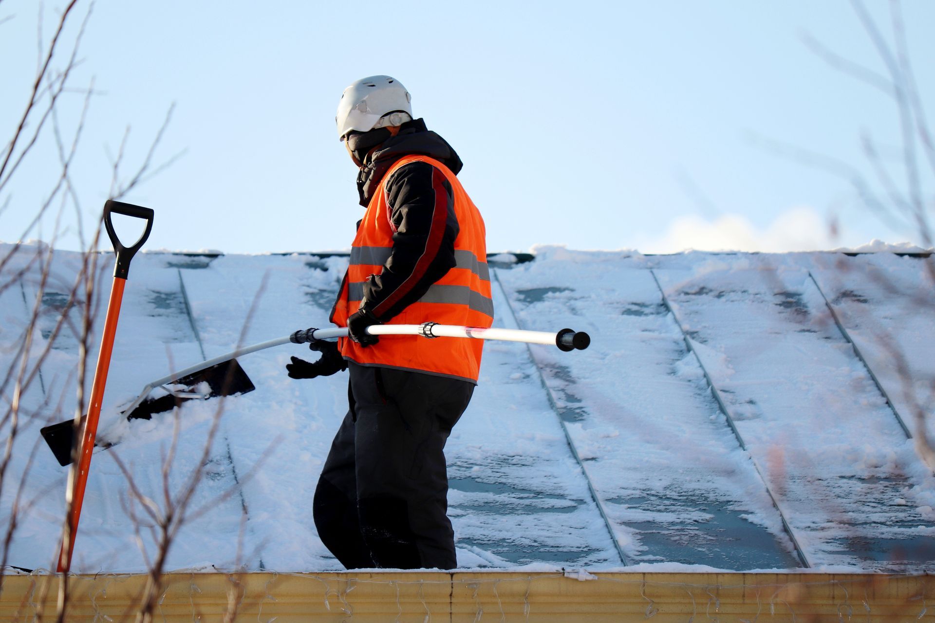 Une personne portant un gilet orange et un casque de sécurité enlève la neige d'un toit à l'aide d'une brosse à neige à long manche.