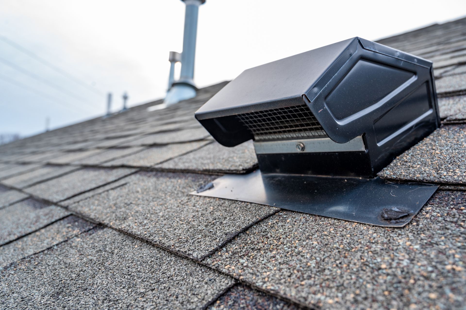 Black roof vent on a gray shingle roof, with a chimney and power lines in the background.