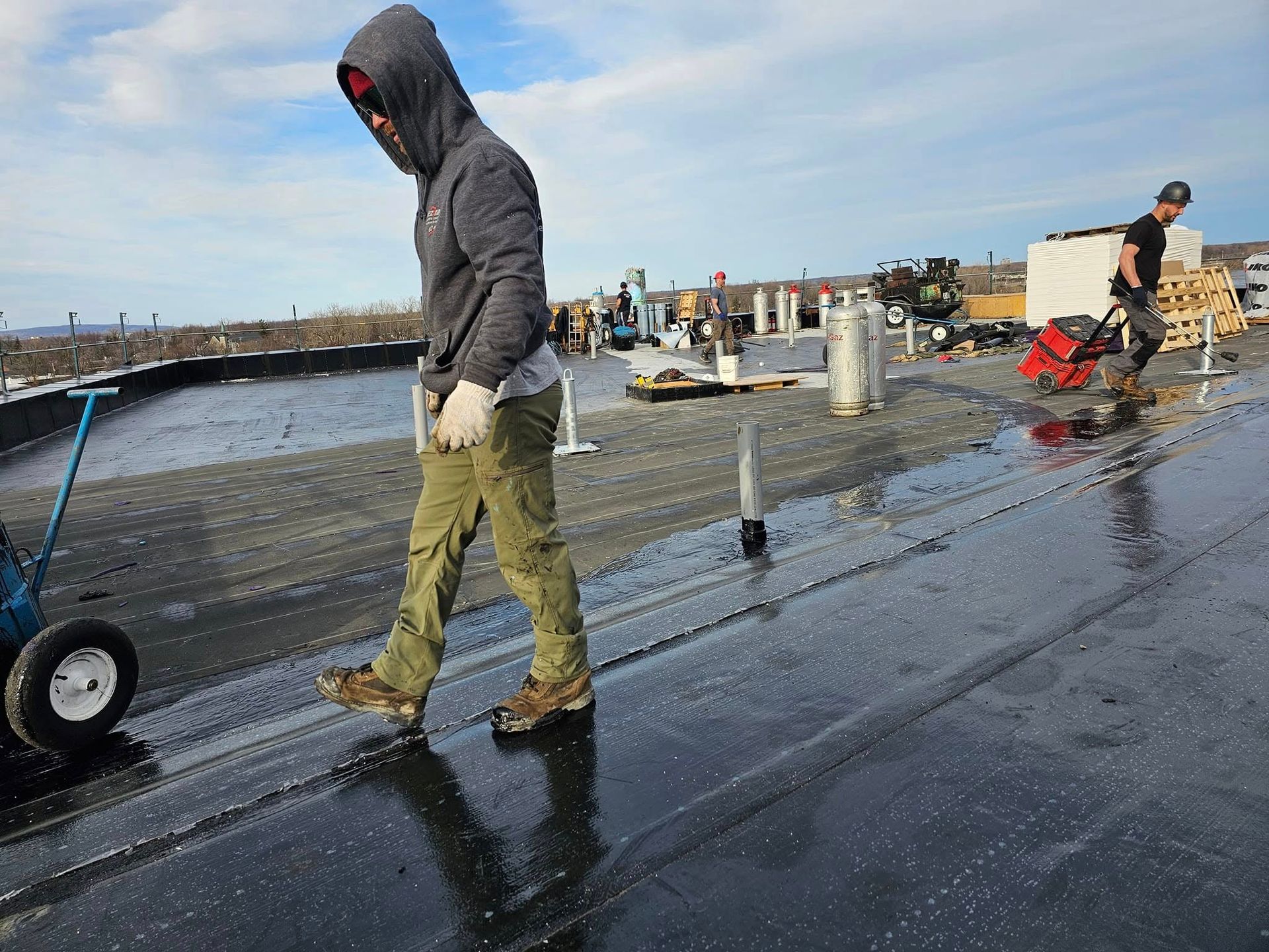 Man in hoodie and work pants on a wet roof, with another person using a machine in the background.