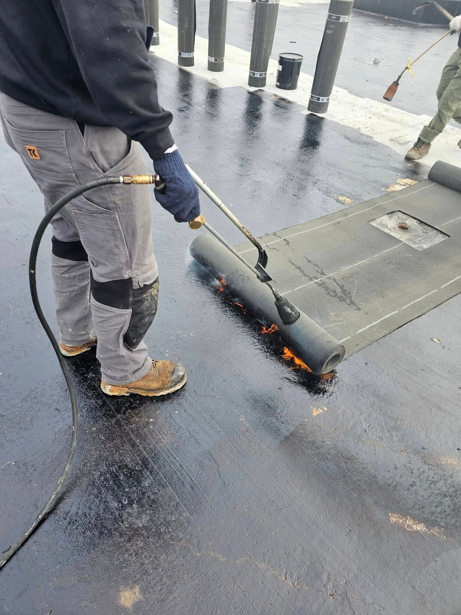 Man using a torch to heat and apply a black roofing material on a dark surface.