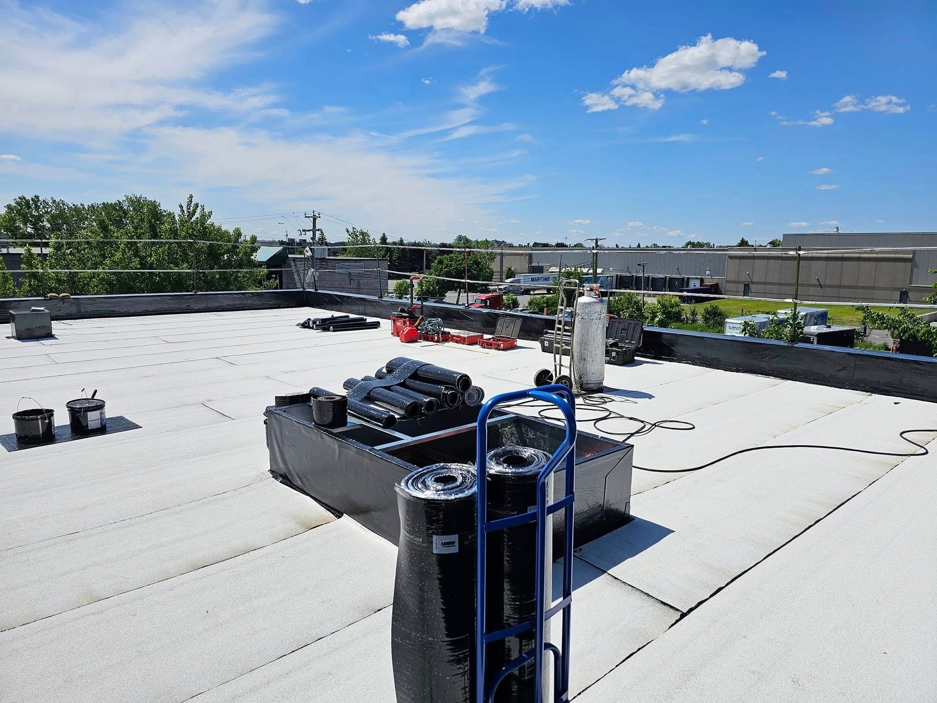 Rooftop scene with equipment, including rolls of roofing material and a propane tank, under a blue sky.