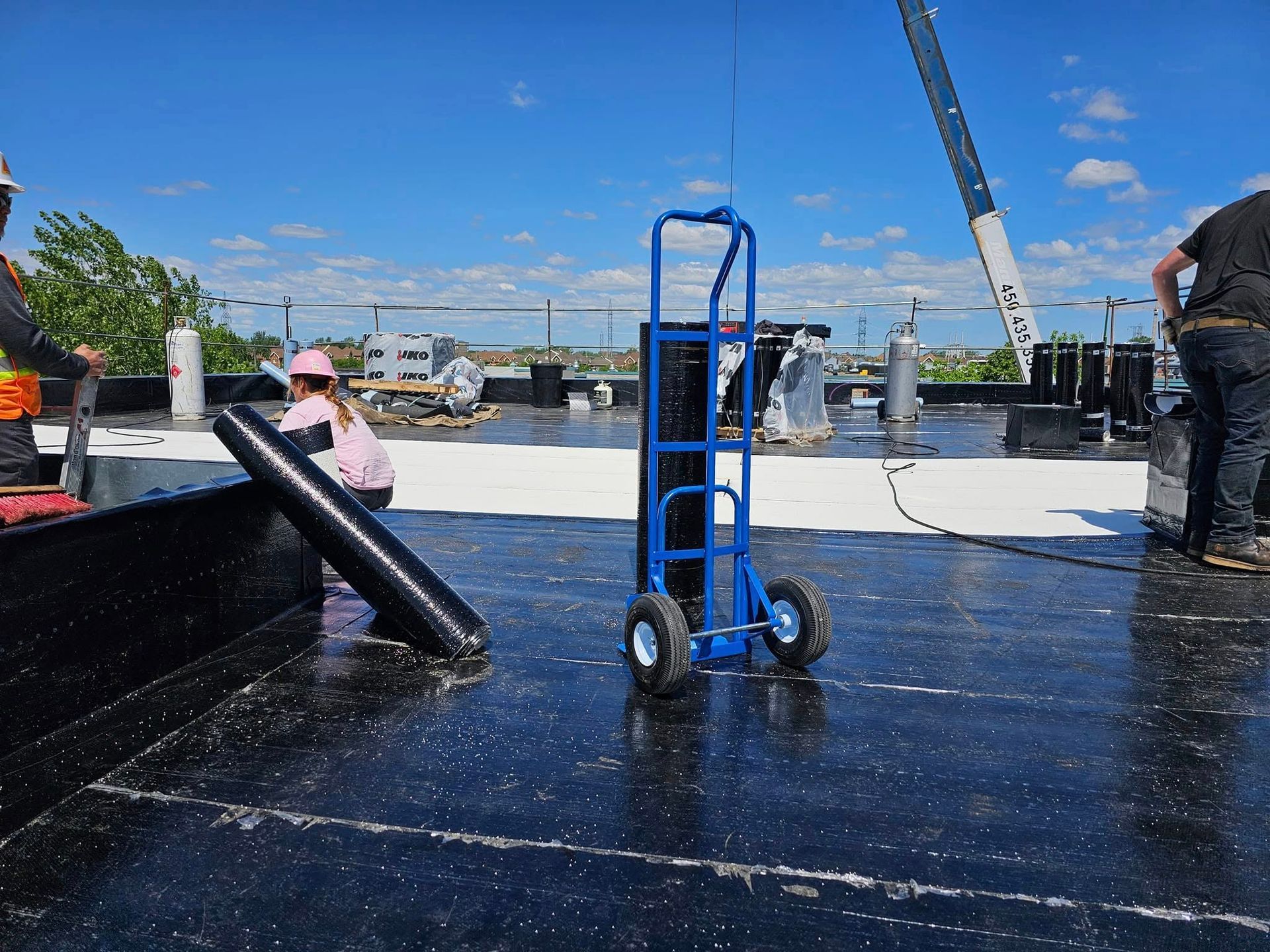 Construction workers on a rooftop. Blue hand truck, black roofing material, crane in the background, sunny day.