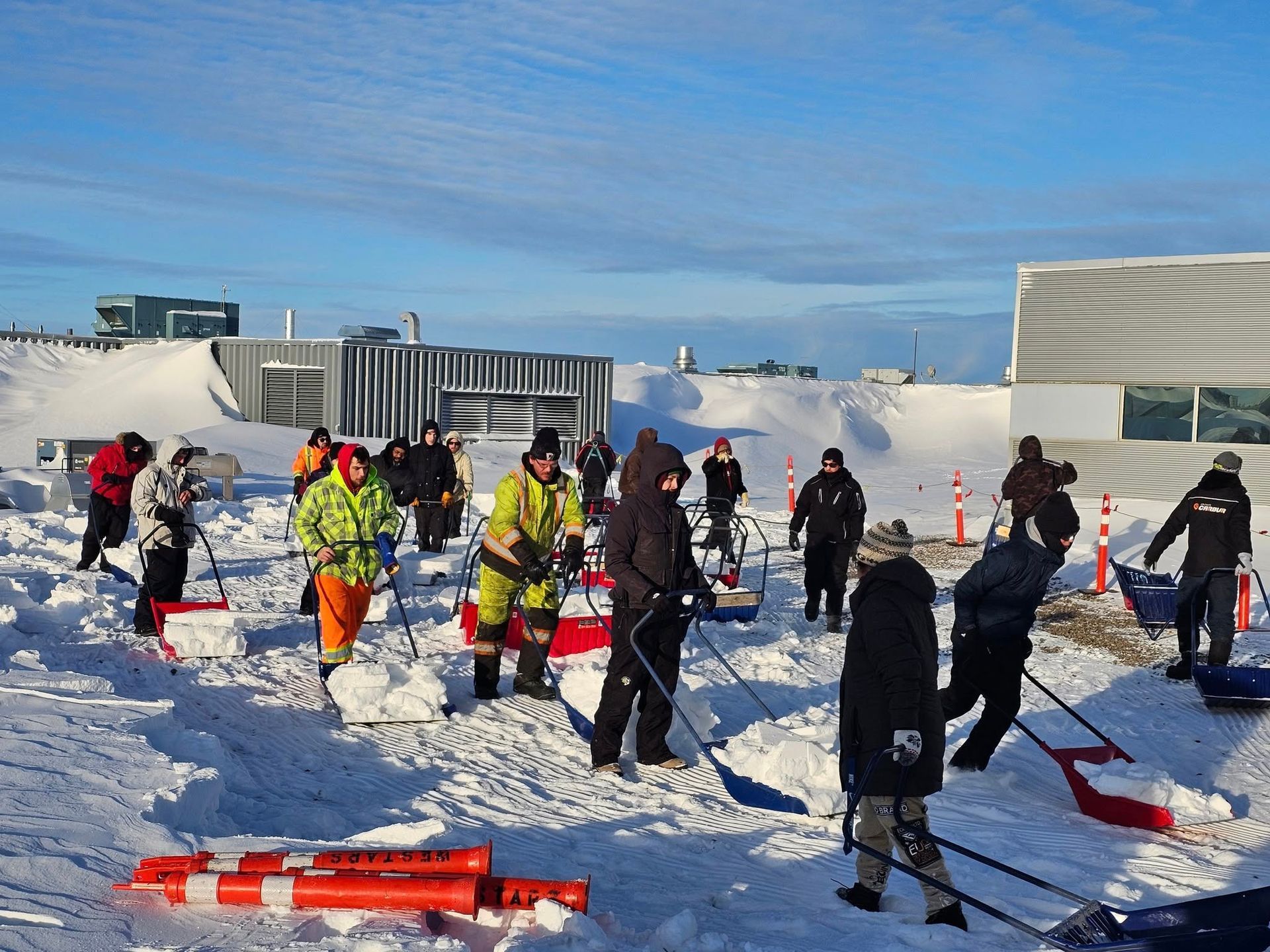 Des gens pelletent la neige à l'extérieur par une journée ensoleillée, près des bâtiments et de l'équipement.