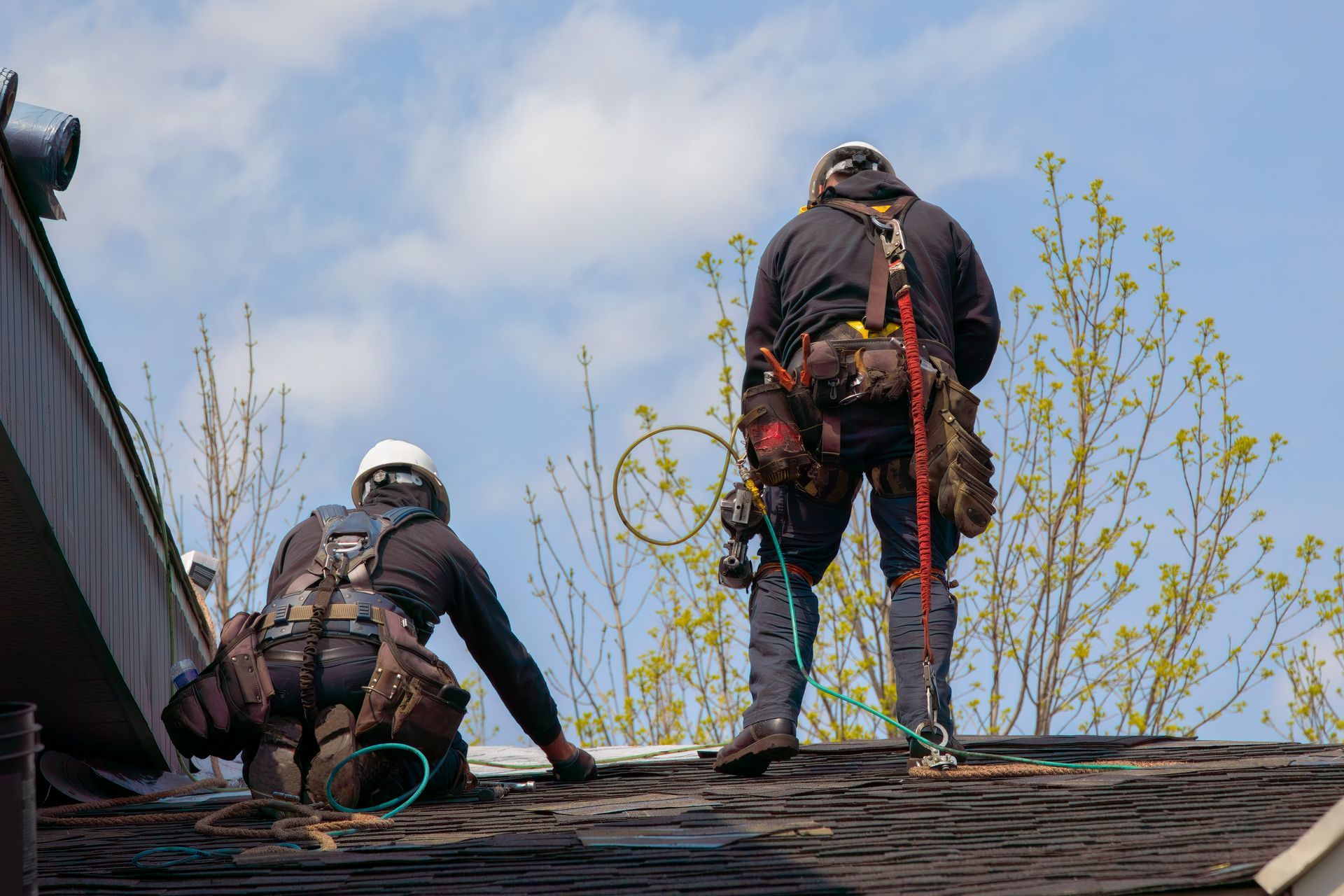 Two roofers wearing safety gear working on a roof with a blue sky background.