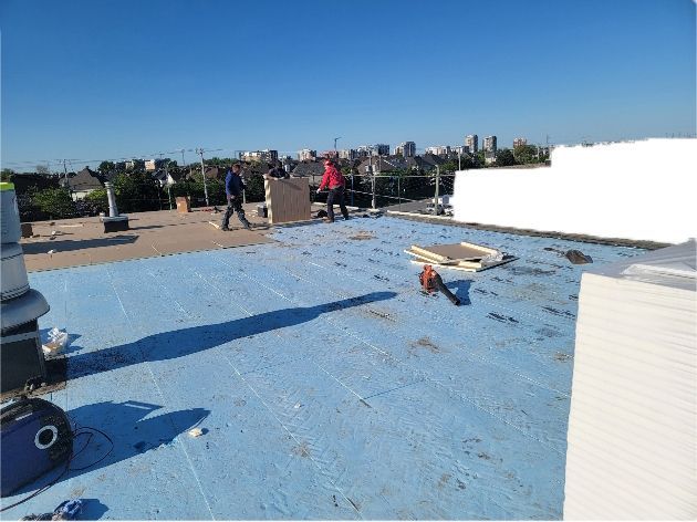 Two roofers working on a blue-covered roof, clearing debris with a blower; city skyline in background.