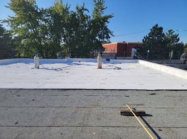 Toit plat recouvert de blanc, deux tuyaux et un bâtiment en arrière-plan. Ciel bleu.