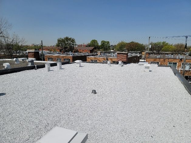Flat, white-graveled rooftop with brick buildings and trees in the background under a blue sky.