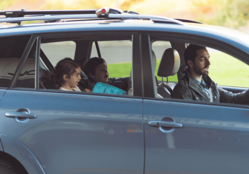 Three people ride in a blue car with a roof rack, seen from the side.