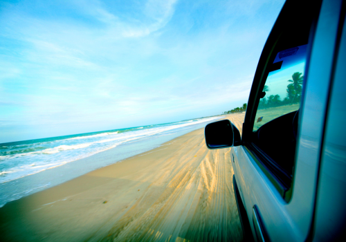 View from a moving car driving along a sandy beach beside blue ocean and sky