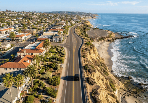 Aerial view of a coastal highway winding along the ocean with homes on one side and rugged cliffs overlooking the shoreline.