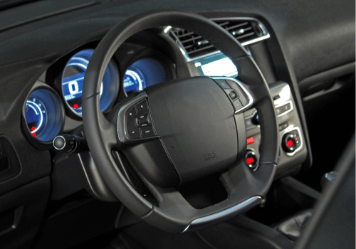 Car interior with steering wheel and illuminated dashboard gauges, viewed from the driver’s seat