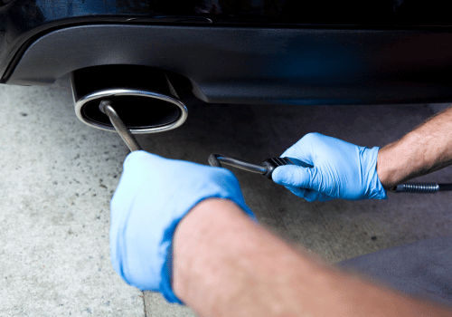 Mechanic in blue gloves using a wrench to tighten a car’s exhaust pipe underneath the bumper