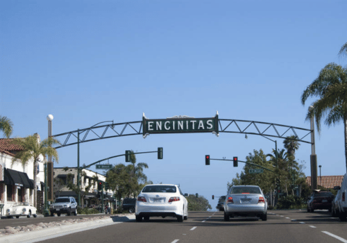 Cars driving under an Encinitas archway on a sunny street with palm trees and traffic lights.