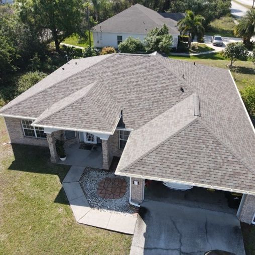 An aerial view of a single-story brick house with a gray shingled roof, a front porch, and a garage in a suburban area.